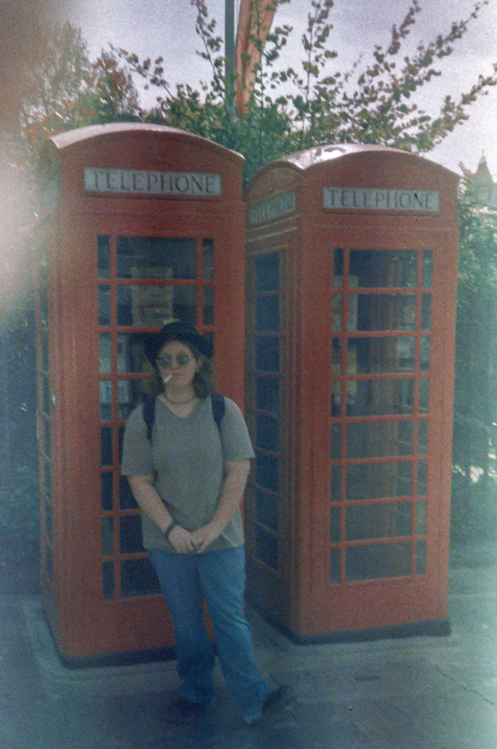 a photo of myself at age 22, wearing a bowler hat and sunglasses, standing in front of 2 red British telephone boxes. i have a cigarette sticking out of my mouth. the image is faded, as it was taken on film